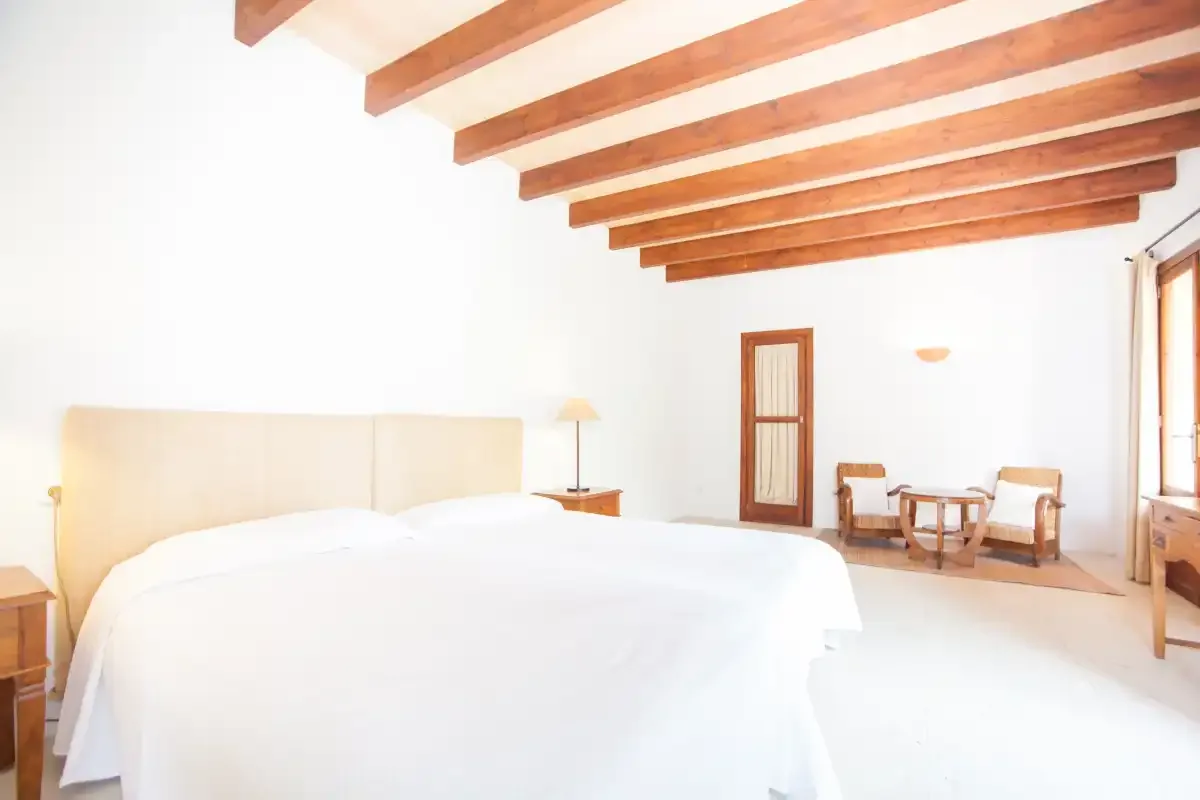 Un dormitorio luminoso con una cama grande, vigas vistas y una pequeña zona de estar.
