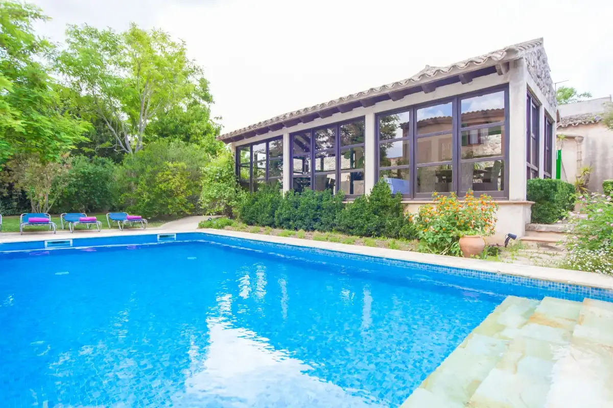 Una piscina exterior está junto a un patio acristalado y vegetación alrededor.