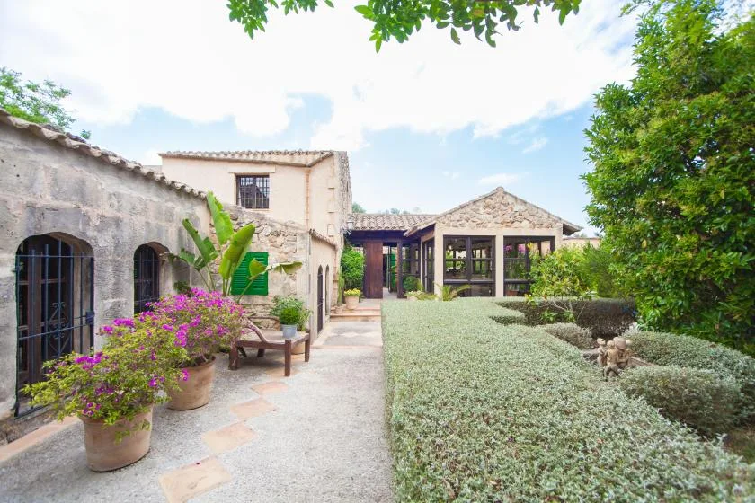 Un patio ajardinado con un camino de piedra, plantas con flores y una zona de estar cubierta.