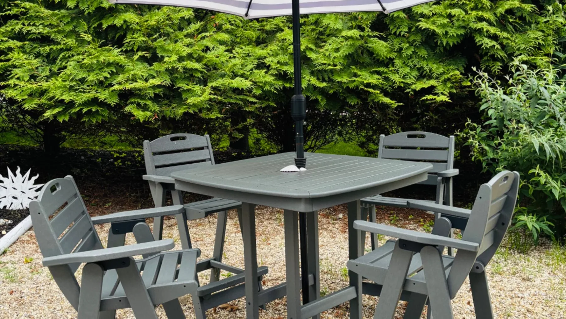 Una mesa gris de patio con asientos está bajo una sombrilla, rodeada de vegetación.