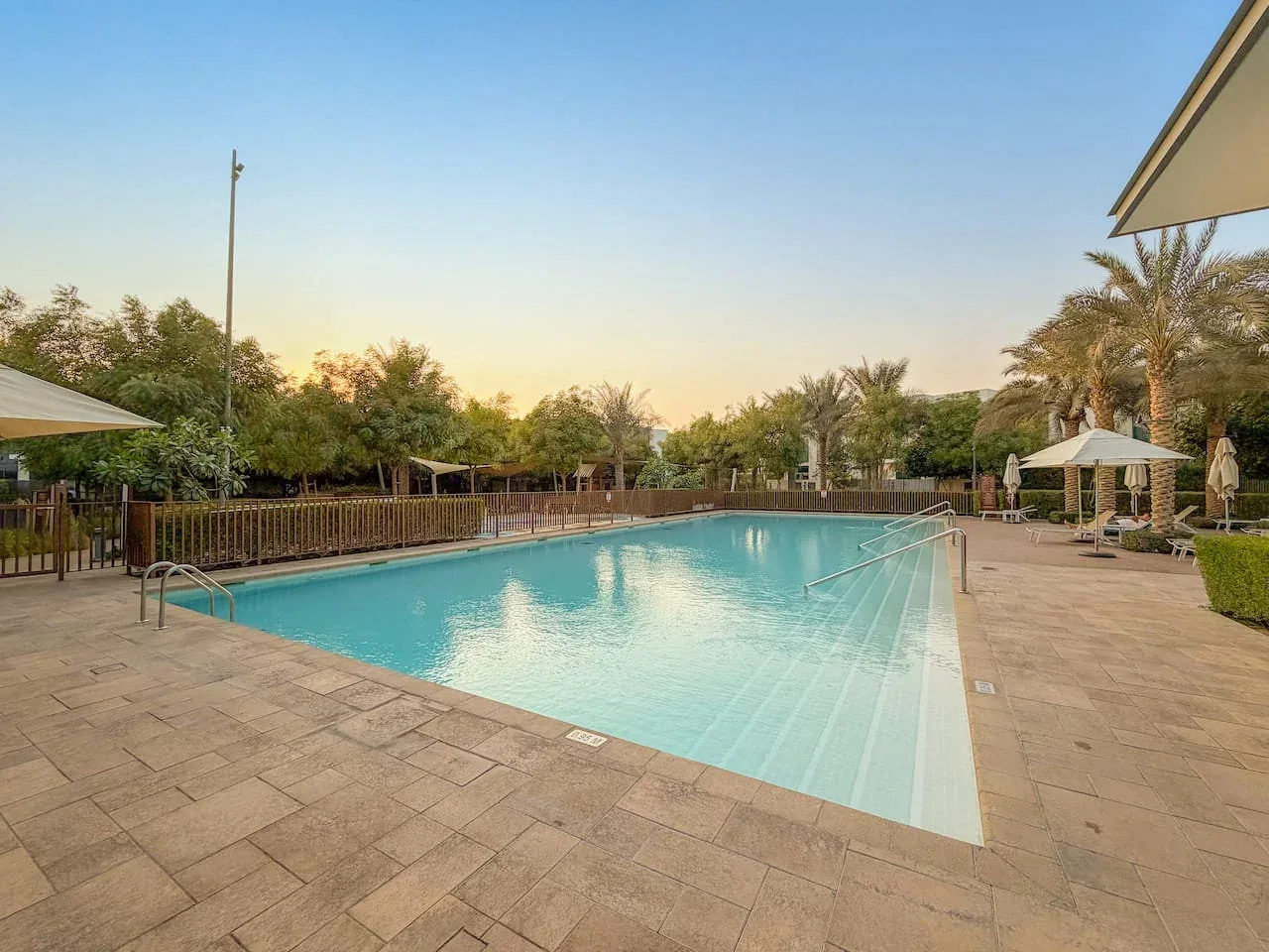 Una piscina al aire libre está rodeada por una terraza pavimentada, árboles y sombrillas.