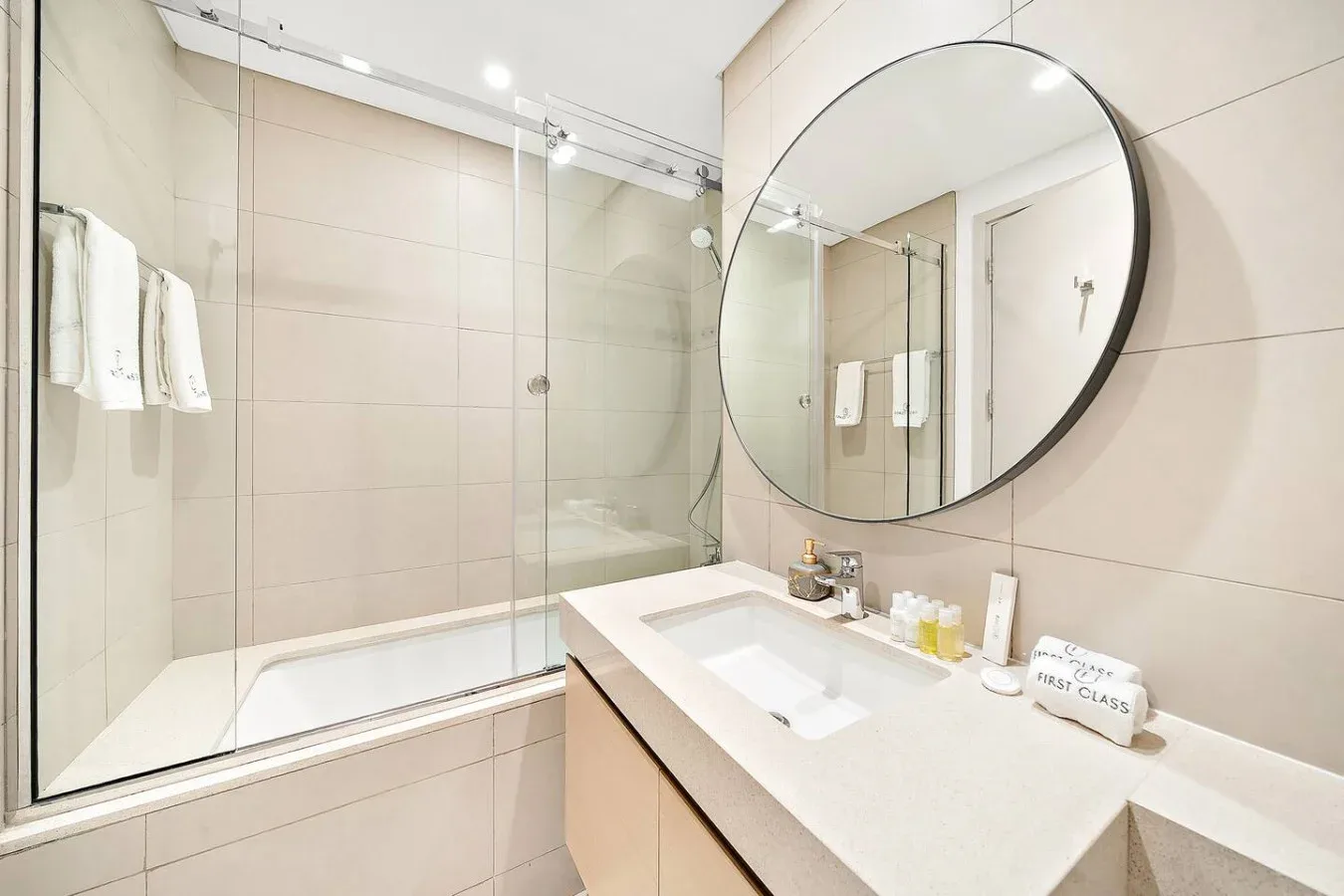 Un baño alicatado con ducha de vidrio, lavabo y un gran espejo redondo.