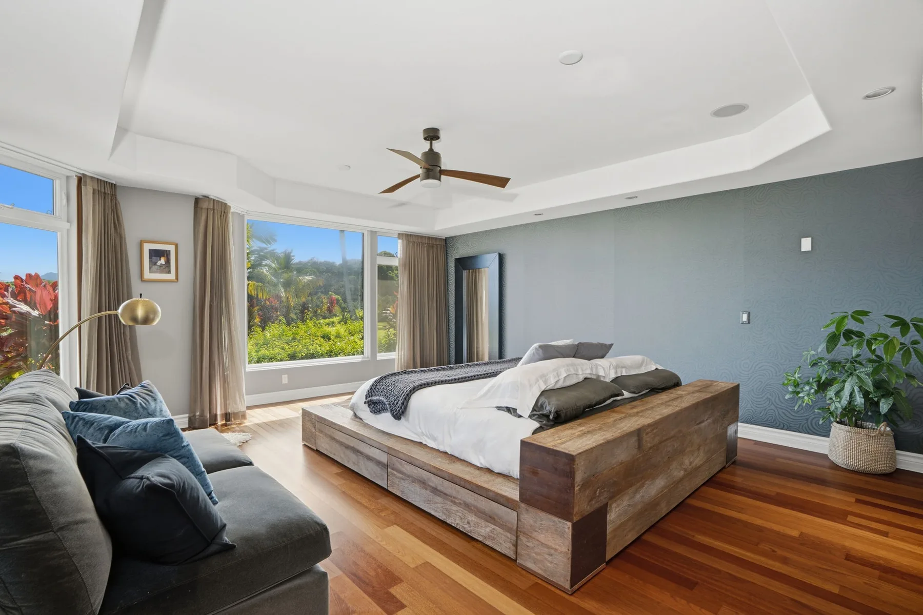 A spacious bedroom with a wooden bed, gray seating, and large windows overlooking greenery.