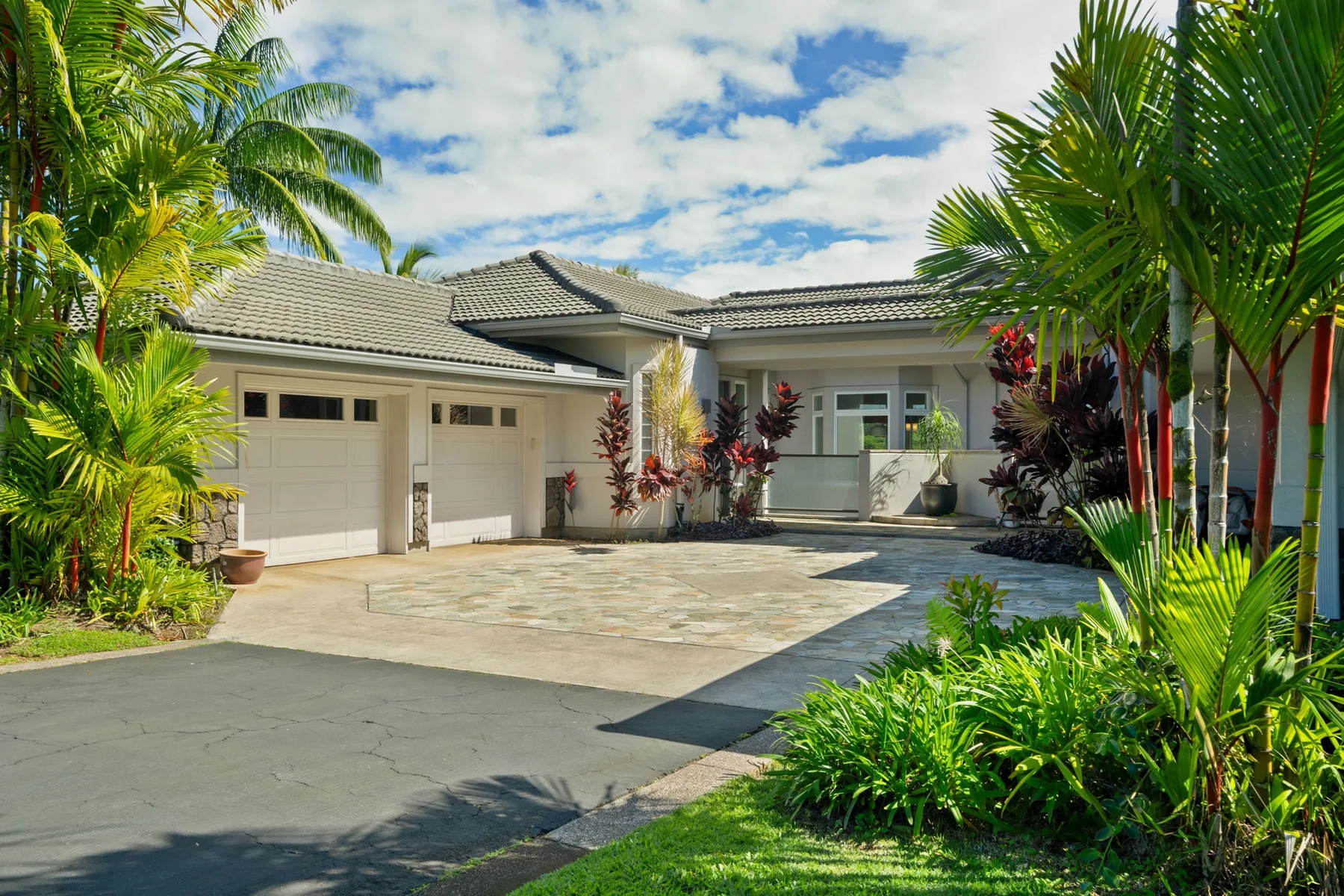A house exterior with garage doors and a paved driveway framed by tropical plants.