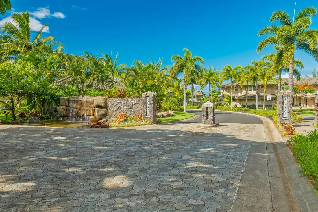 A paved entrance drive curves past a stone sign amid palm trees and landscaping.