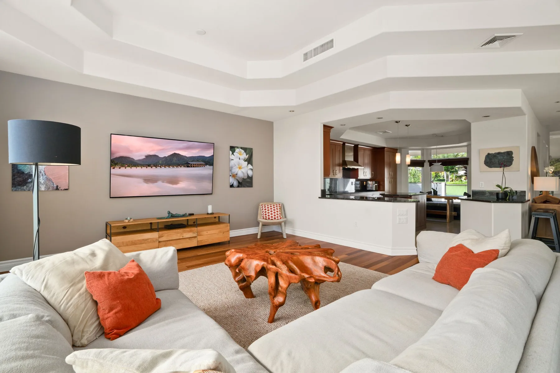 A bright living room with light-colored seating, a wooden coffee table, and a kitchen beyond.