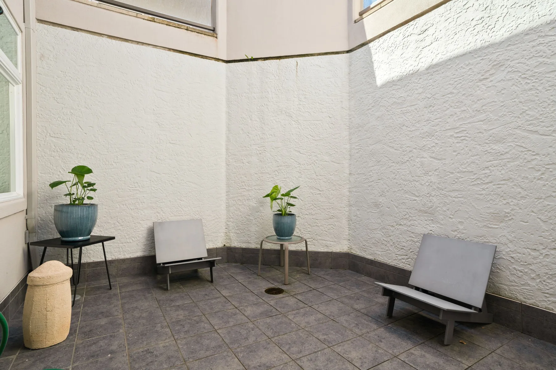 A small open-air patio with tiled flooring, seating, and potted plants.