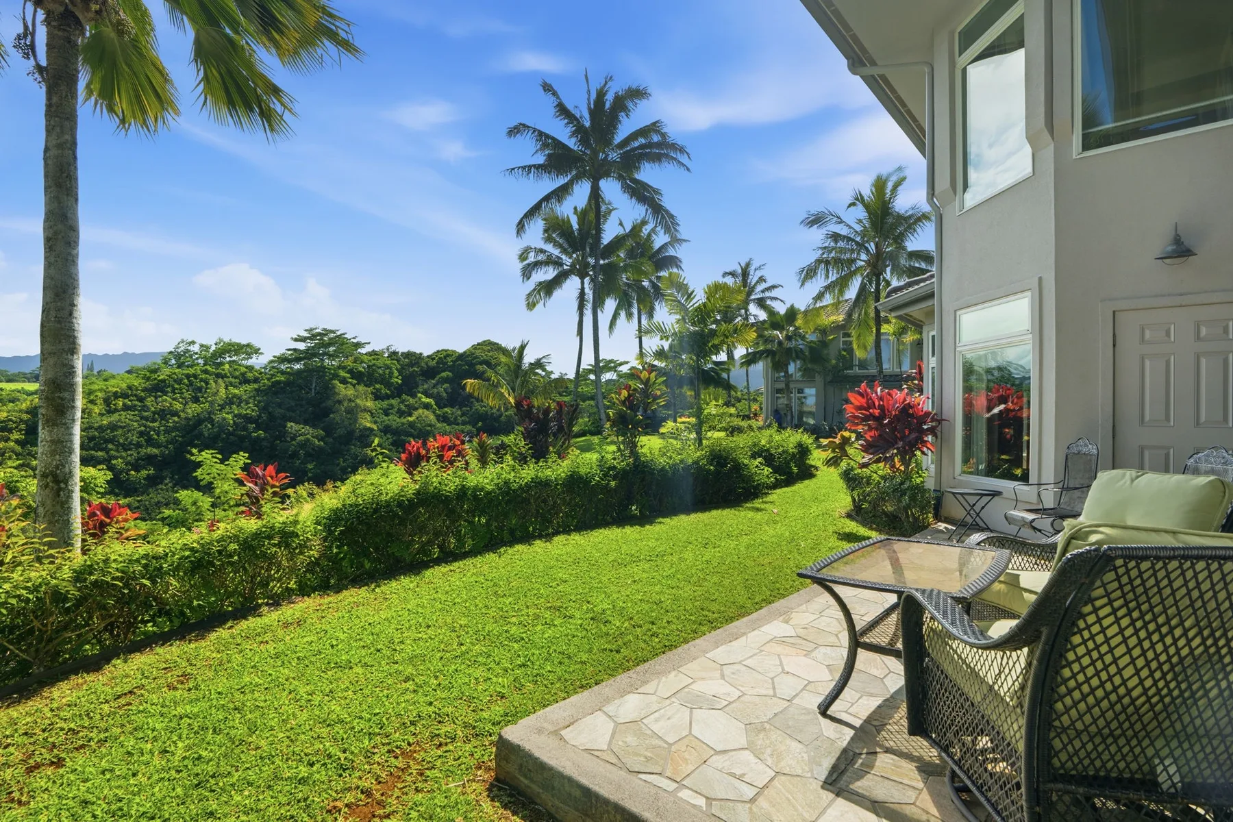 A patio beside a house overlooks a green lawn, tropical plants, and palm trees.