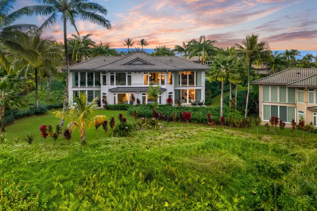 A large house stands amid tropical greenery beneath a colorful sunset sky.