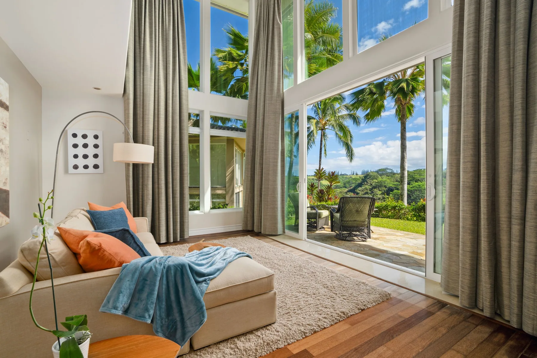 A bright living room has tall windows overlooking palm trees and a garden.