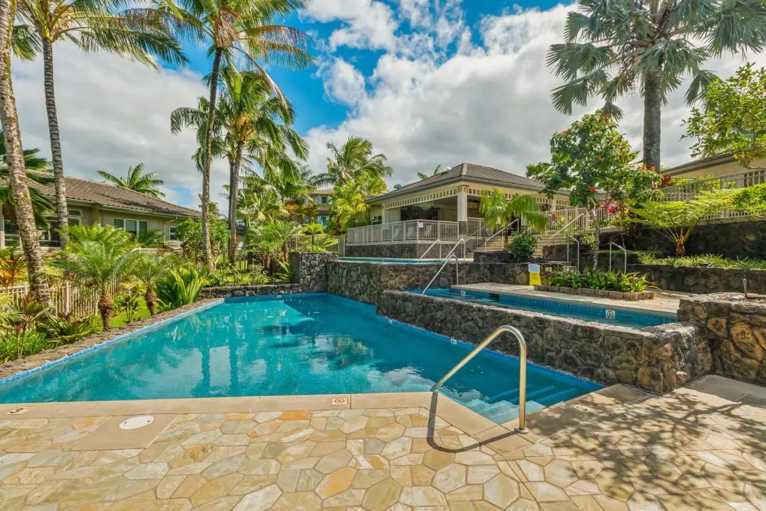 A swimming pool sits beside a stone wall, surrounded by tropical landscaping.