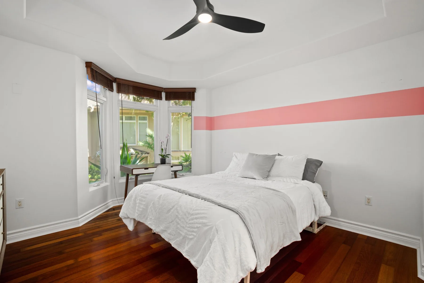 A bright bedroom has a white bed beside large windows and a small desk.