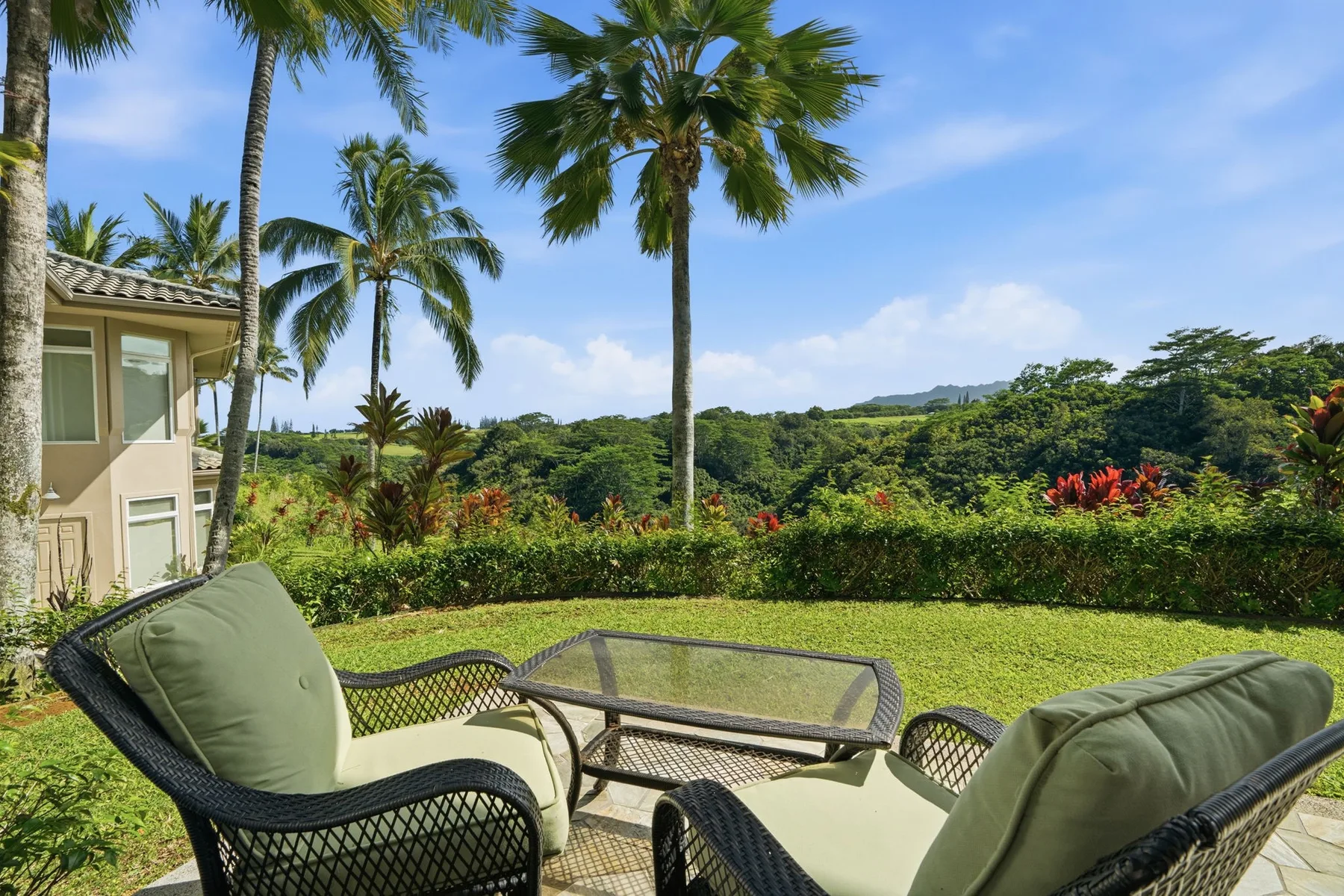 An outdoor patio with cushioned seating overlooks a green lawn and tropical trees.
