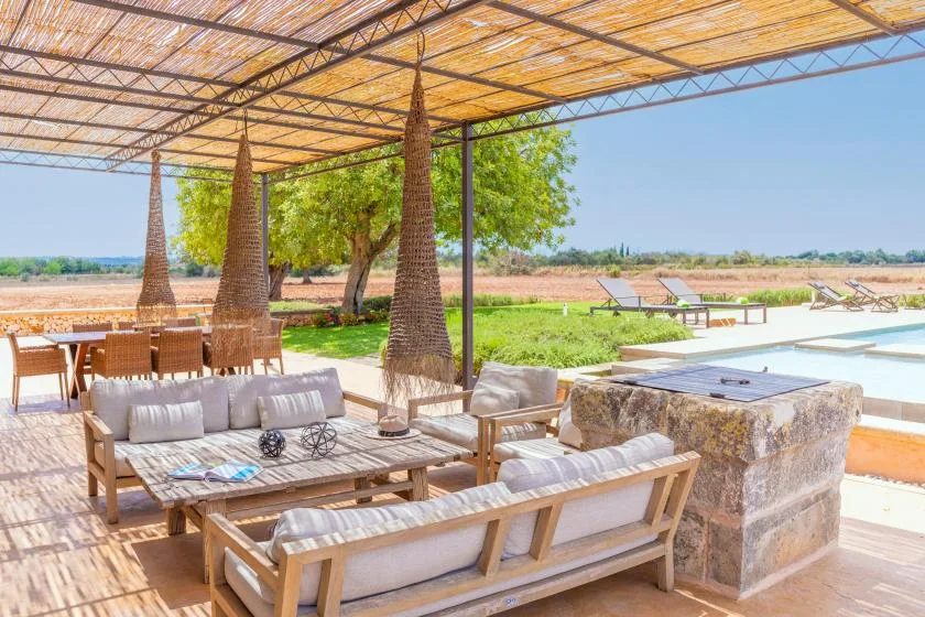 Una sala exterior sombreada con asientos acolchados y una mesa de piedra da a un paisaje abierto.