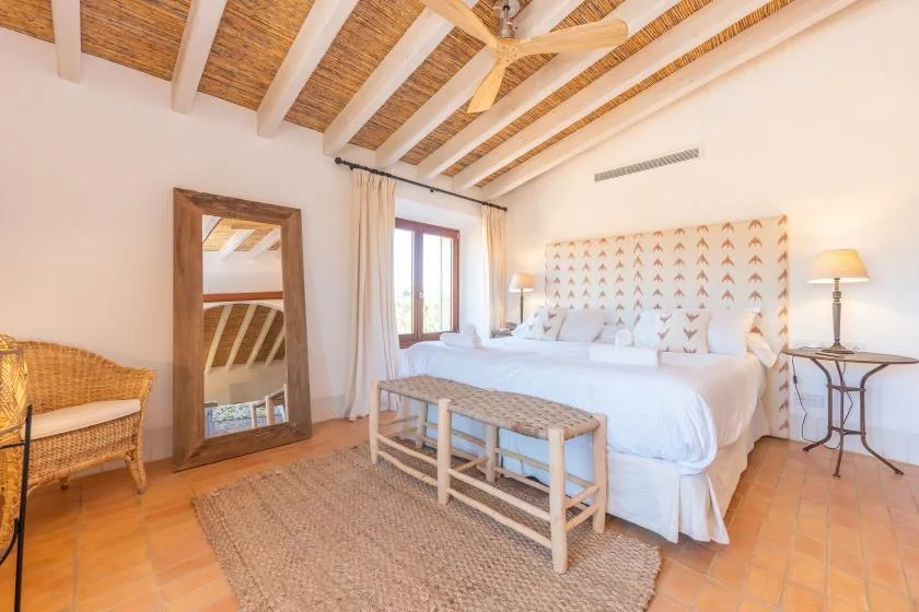 Un dormitorio luminoso con una cama blanca, un banco tejido y vigas expuestas en el techo.