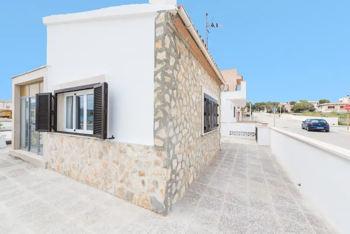 A white house with stone accents faces a paved walkway and low wall.