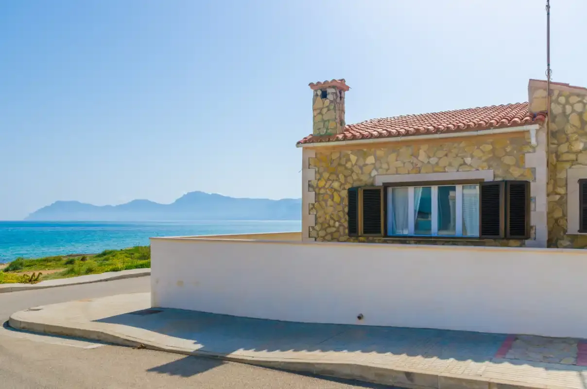 A stone house beside a coastal road, with the sea and mountains in the background.