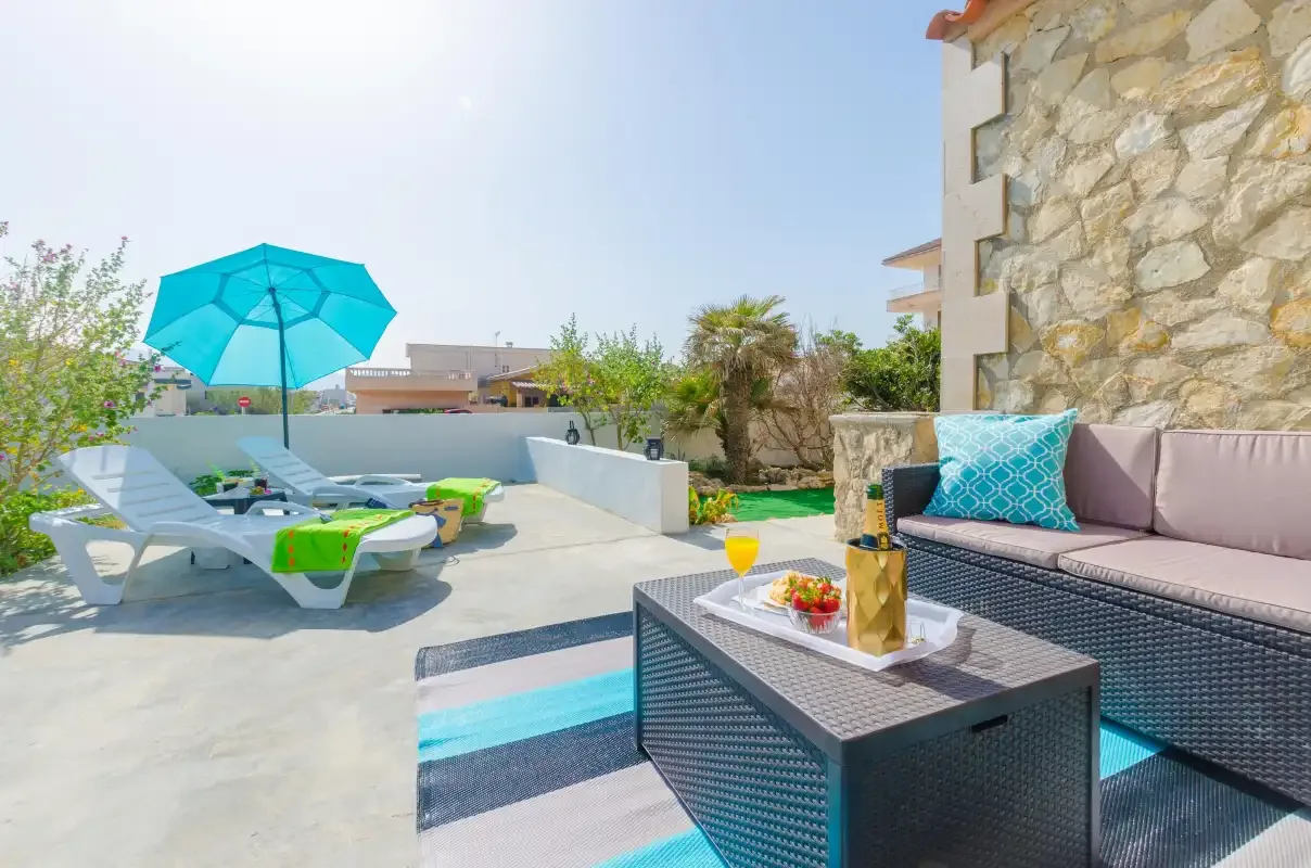 A sunny outdoor patio has lounge seating, a turquoise umbrella, and a low table.