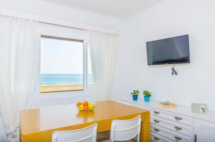 A bright room with a wooden table, white seating, and a television beside a sea view.