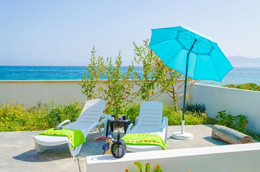 A seaside patio with lounge seating, a turquoise umbrella, and ocean views.