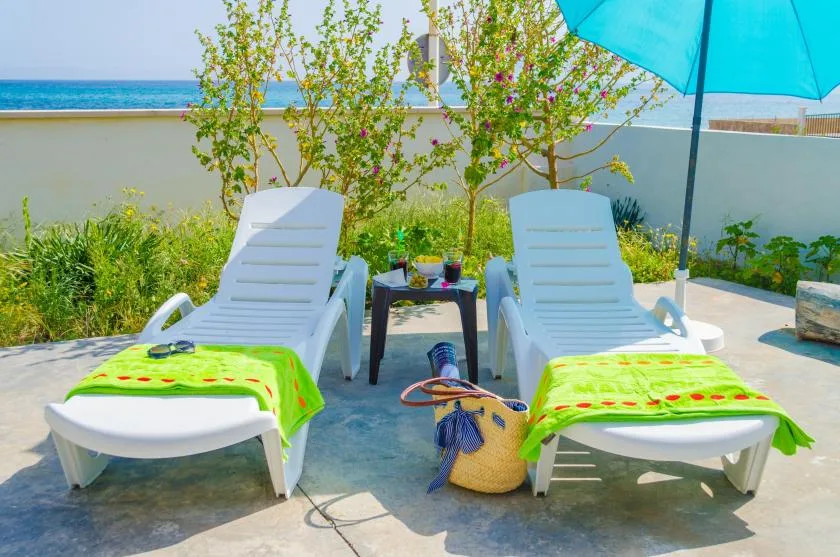 Two lounge chairs and a small table sit on a sunny patio beside the sea.