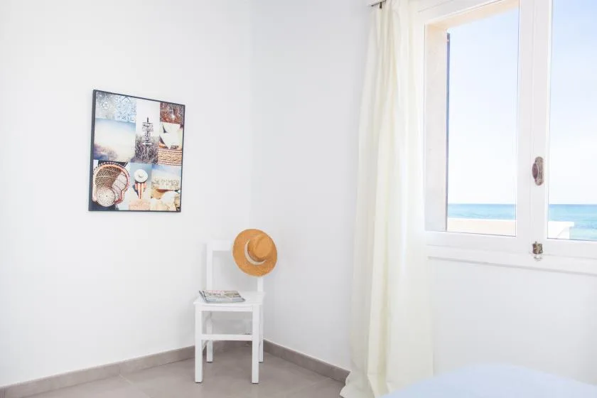 A bright bedroom with a small white side table beside a window overlooking the sea.