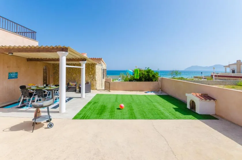A sunny outdoor patio has a green lawn, shaded seating, and a view of the sea.