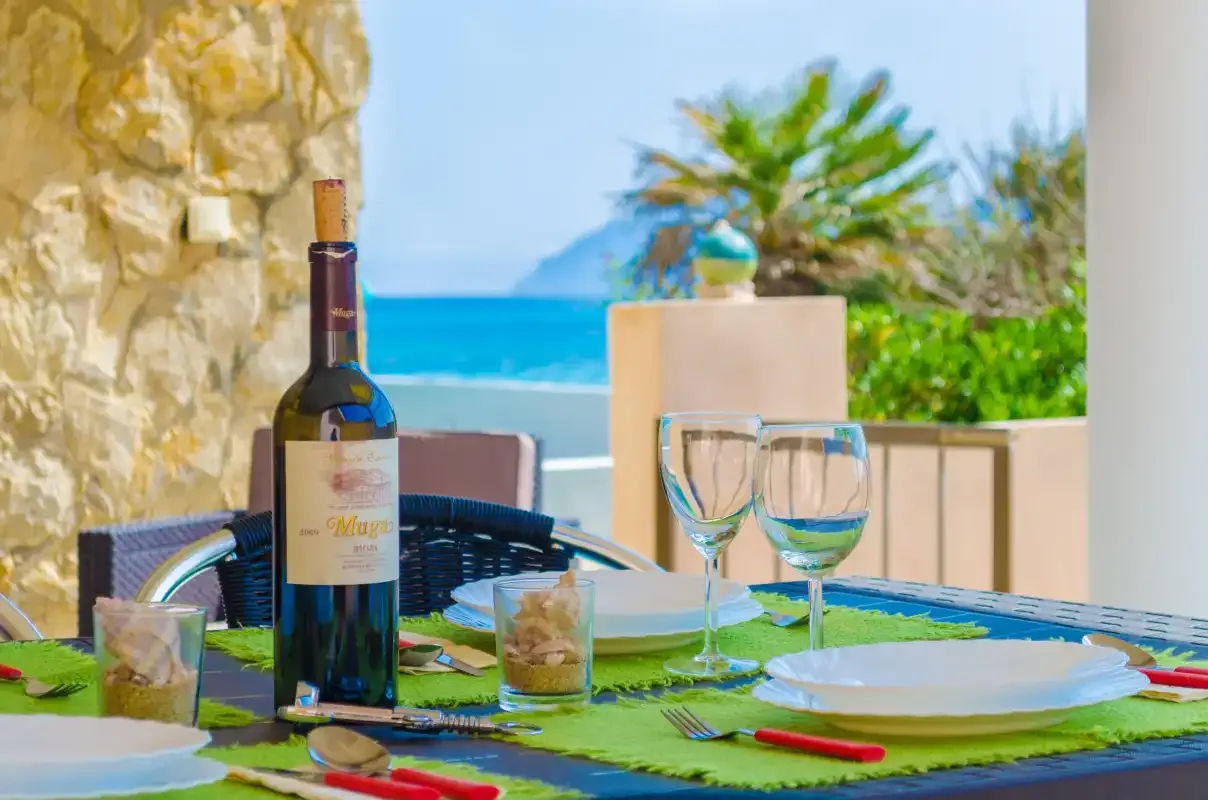 A seaside dining table is set with plates and wine glasses beside a stone wall.