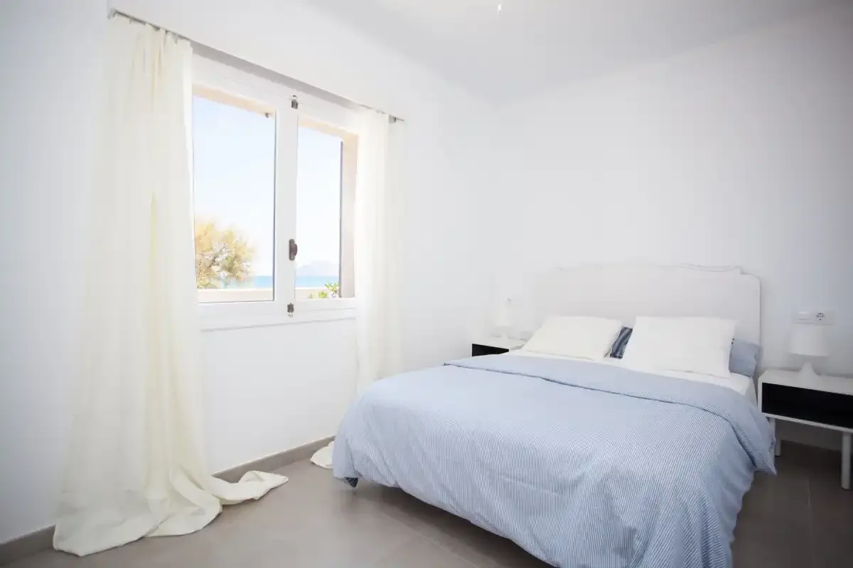 A bright bedroom with a neatly made bed and a window overlooking the sea.