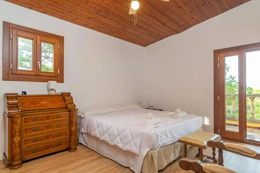 A bedroom with a bed, wooden dresser, and windows opening onto greenery.