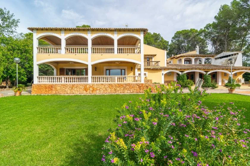 A large house with arched balconies stands behind a green lawn and flowering shrubs.