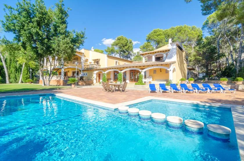 A swimming pool borders a sunny patio in front of a large house.