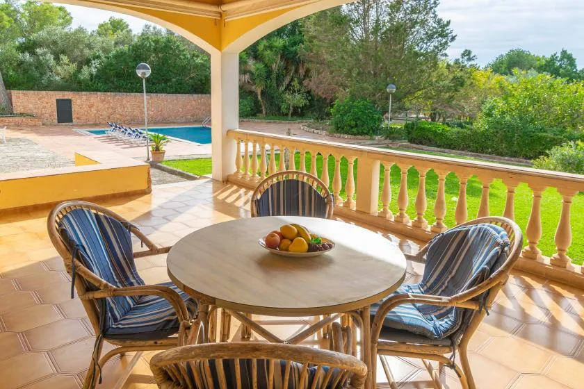 A covered patio has a round table and seating overlooking a pool and garden.
