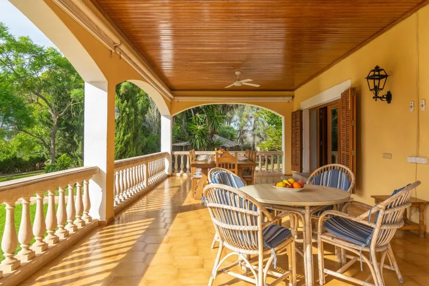 A covered veranda with a dining table and chairs overlooking green trees.
