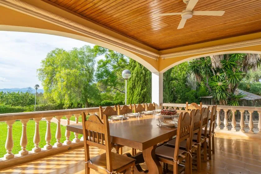 A covered outdoor dining area with a wooden table and chairs overlooking greenery.
