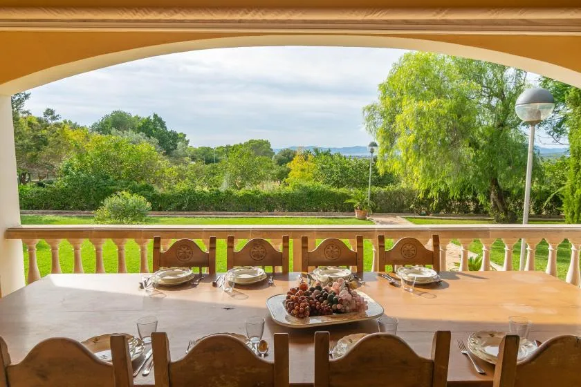 A dining table set with food sits on a covered terrace overlooking a leafy garden.