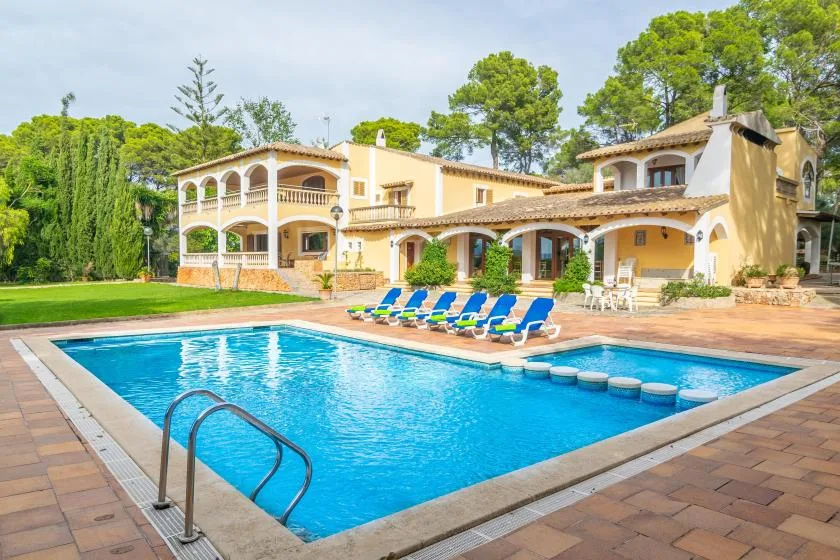 A swimming pool sits in front of a large villa, with lounge chairs beside the water.
