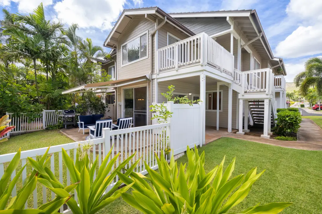 Una casa de dos plantas con balcones blancos, césped y plantas tropicales.