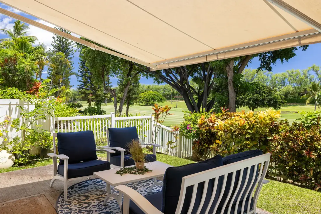 Un patio al aire libre con asientos acolchados, una mesa y una baranda blanca da a un jardín.