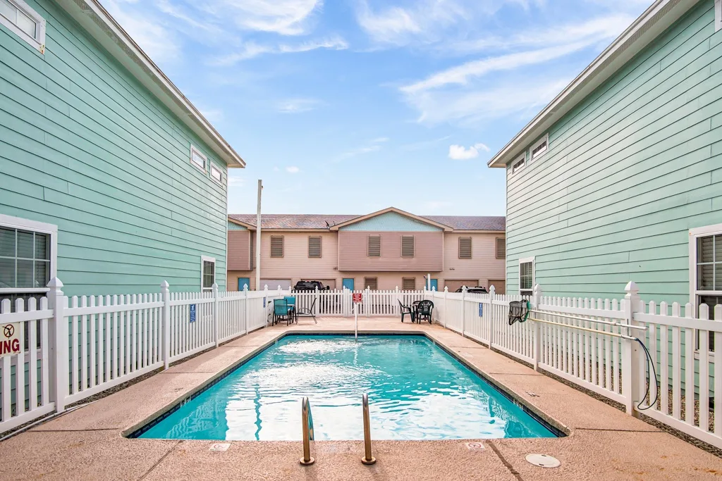 An outdoor swimming pool sits between green-sided buildings under a blue sky.