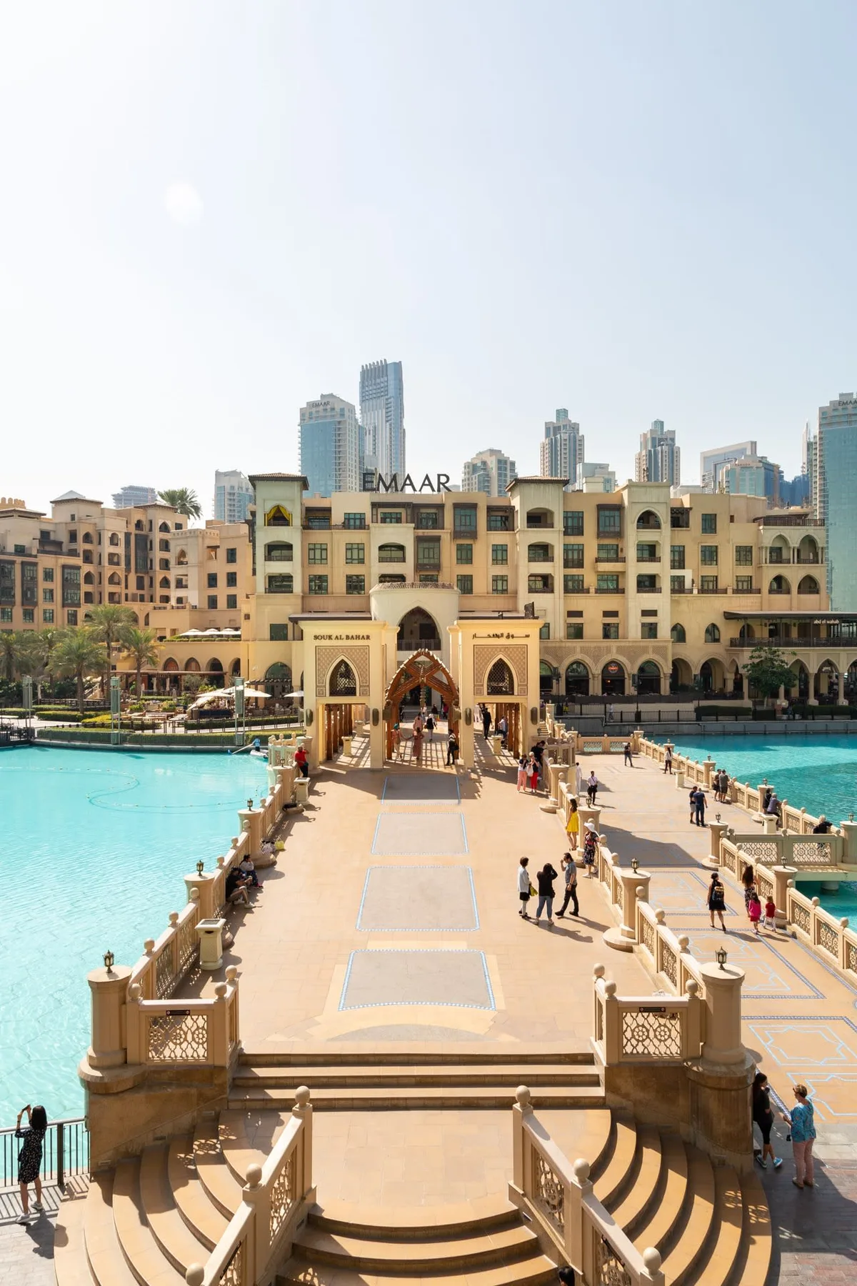 A broad walkway crosses a pool toward an ornate building, with city towers behind it.