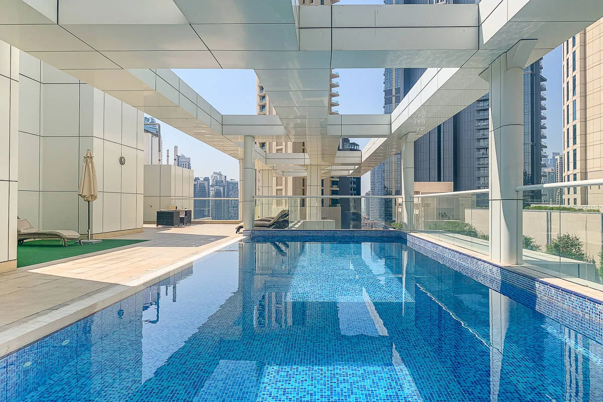 A rooftop swimming pool sits beneath a glass canopy, with city buildings visible beyond.