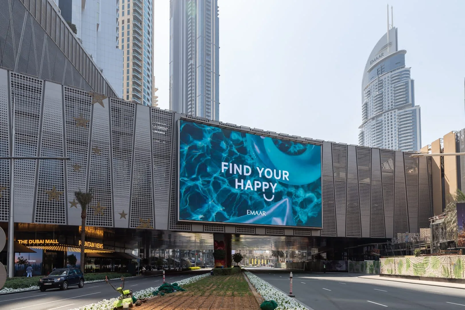 A city street runs beside a large building with a digital billboard and tall towers behind it.