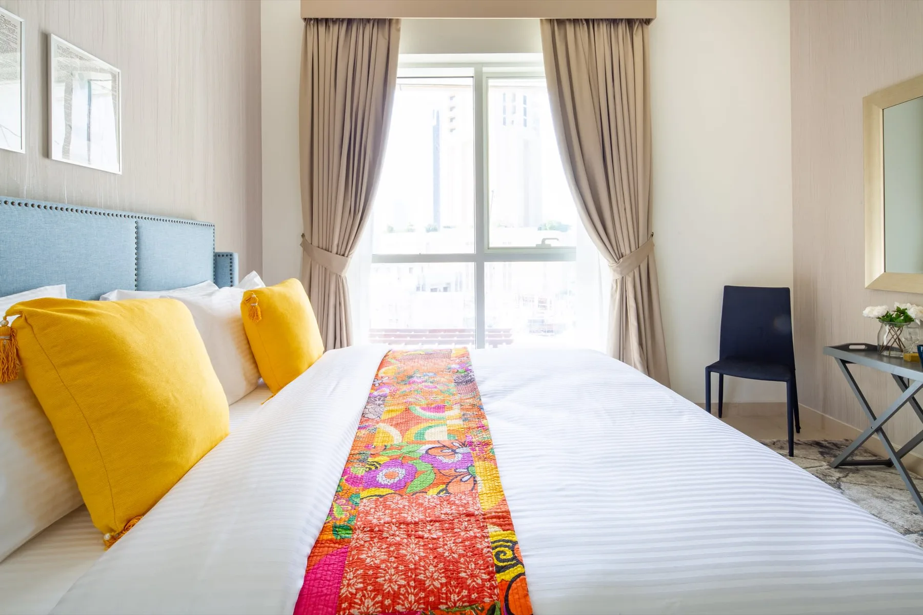 A bright bedroom with a large bed and colorful runner beneath a curtained window.