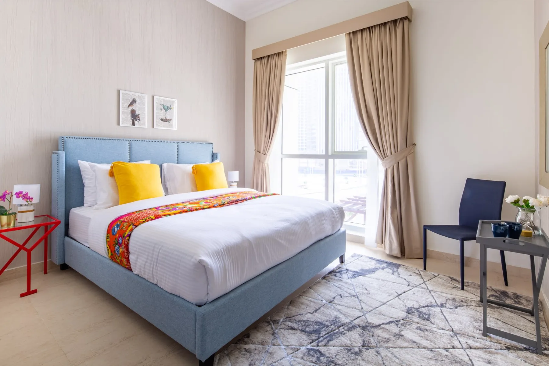 A bedroom with a blue bed, colorful pillows, and a large curtained window.