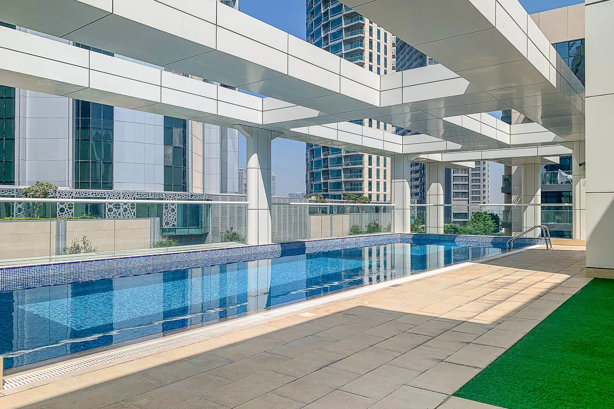 A swimming pool sits beside a paved deck beneath a modern building canopy.