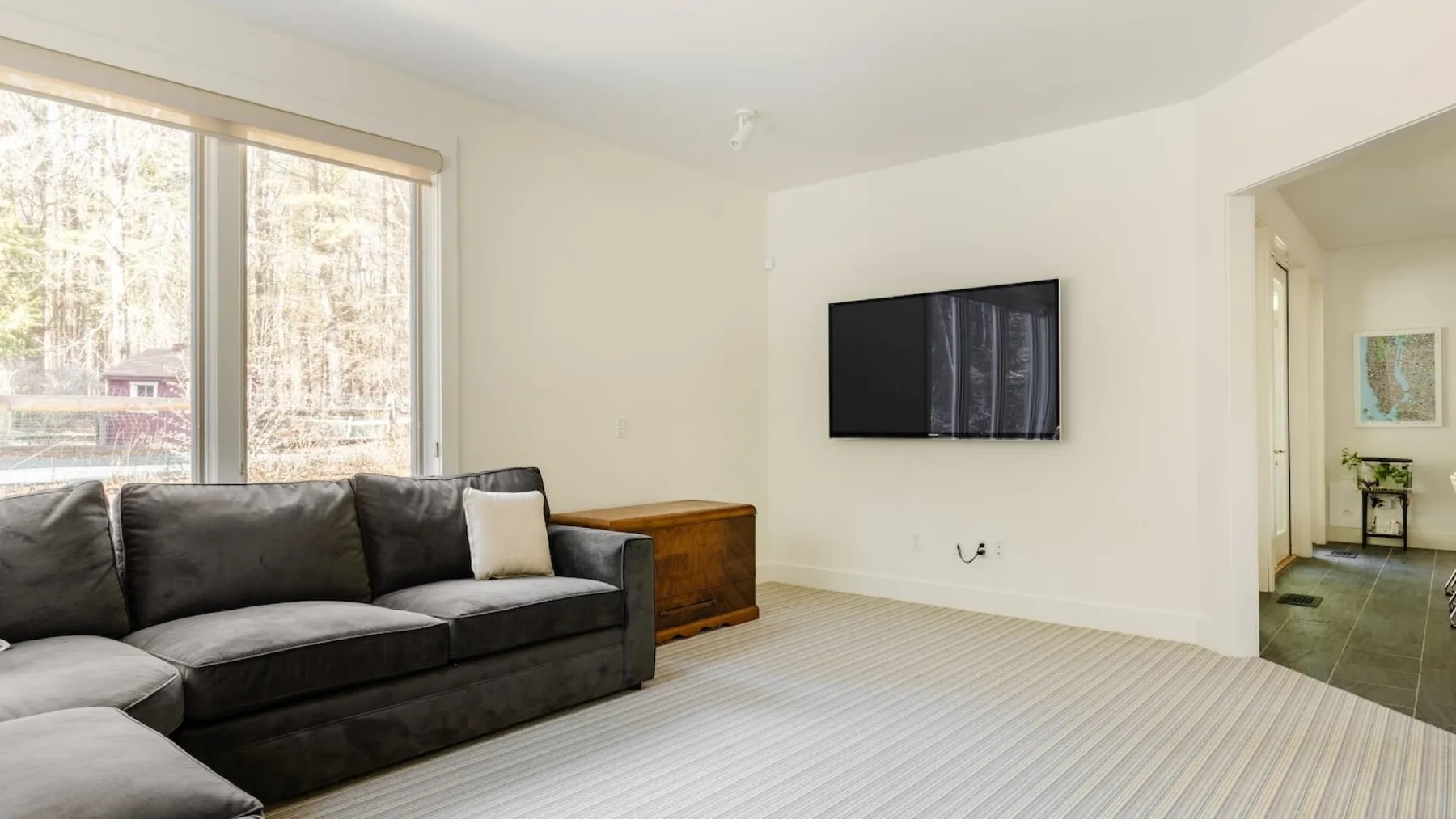 Una sala de estar con un sofá seccional oscuro, un televisor en la pared y ventanales.