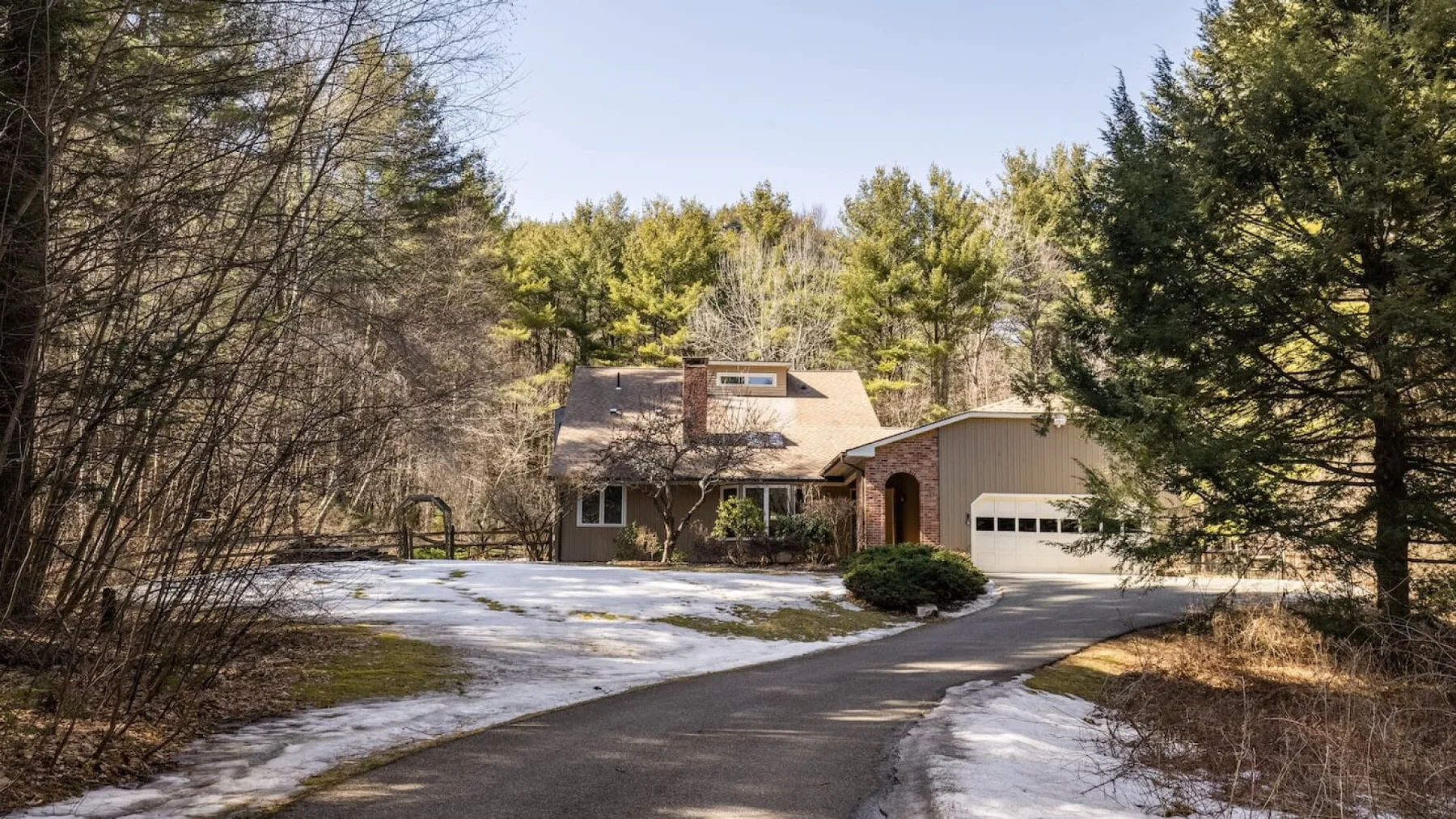 Una casa y un garaje están junto a un camino curvo, con nieve entre los árboles.