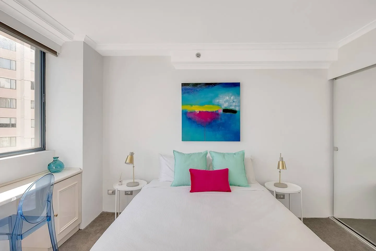 A bright bedroom features a bed with teal pillows and colorful artwork above it.