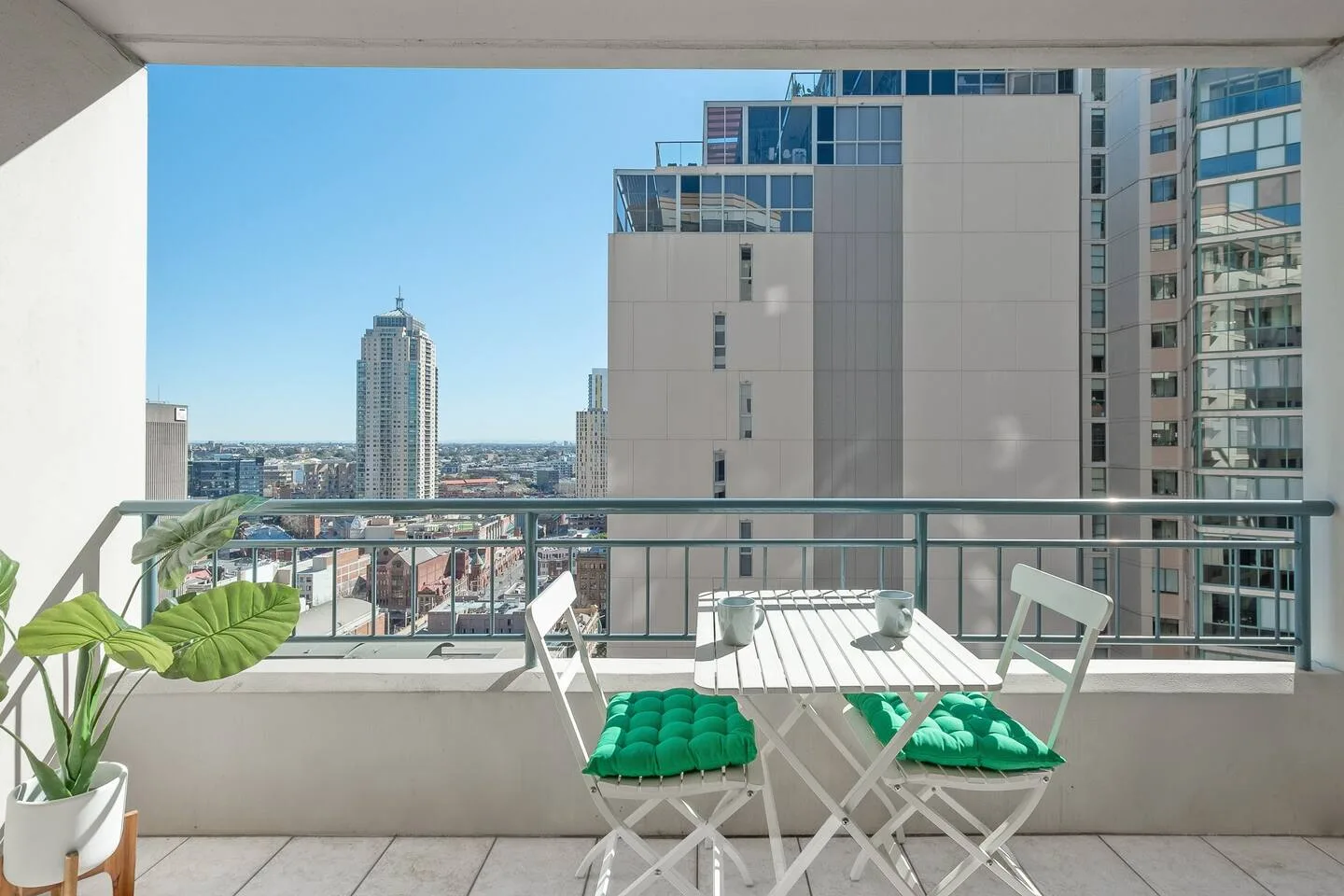 A balcony seating area overlooks nearby high-rise buildings beneath a clear blue sky.